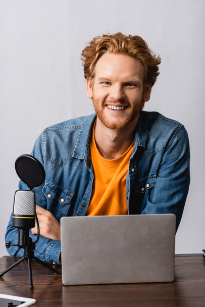 excited radio host in denim shirt looking at camera near laptop and microphone - Foto, Imagen