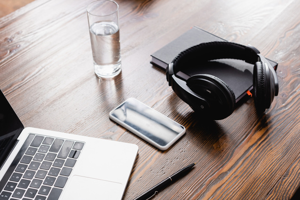 bezdrátová sluchátka, smartphone, notebook, notebook a sklenice vody na kancelářském stole - Fotografie, Obrázek