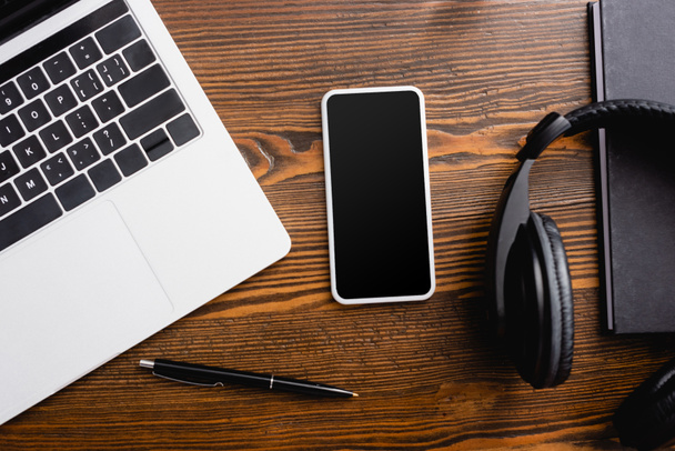 vista superior del teléfono inteligente con pantalla en blanco, ordenador portátil, auriculares inalámbricos, portátil y pluma en el escritorio de la oficina - Foto, Imagen