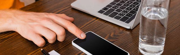 cropped view of teleworker touching smartphone with blank screen near laptop and glass of water, horizontal concept - Photo, Image