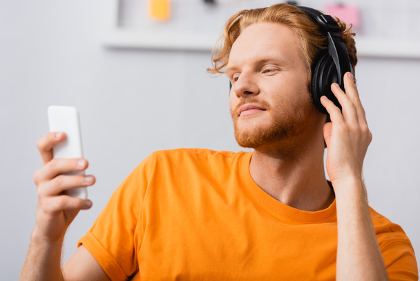 selective focus of dreamy man touching wireless headphones while holding smartphone - Foto, Imagen