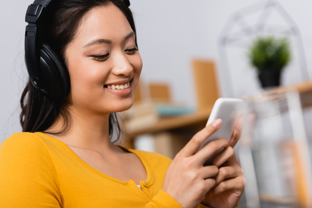 selective focus of brunette asian woman using smartphone while listening podcast in wireless headphones - Valokuva, kuva