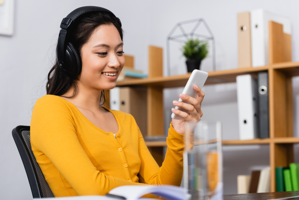 selective focus of brunette asian woman listening podcast in wireless headphones while using smartphone at home - Fotoğraf, Görsel