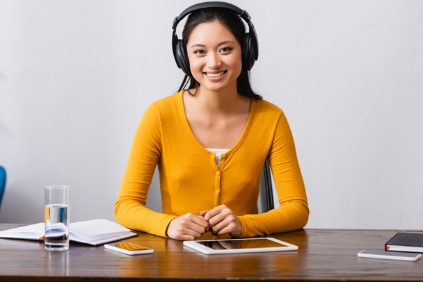 junge asiatische Studentin mit drahtlosen Kopfhörern blickt in die Kamera, während sie am Schreibtisch in der Nähe von Geräten, Notebooks und einem Glas Wasser sitzt - Foto, Bild