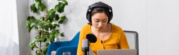 imagen horizontal de joven asiático radio host en auriculares inalámbricos trabajando cerca de micrófono y portátil en estudio - Foto, Imagen