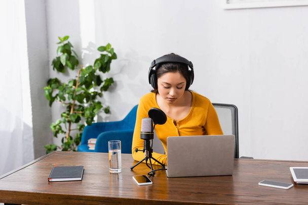 morena asiático emissora usando laptop perto de microfone, vidro de água e gadgets no local de trabalho - Foto, Imagem