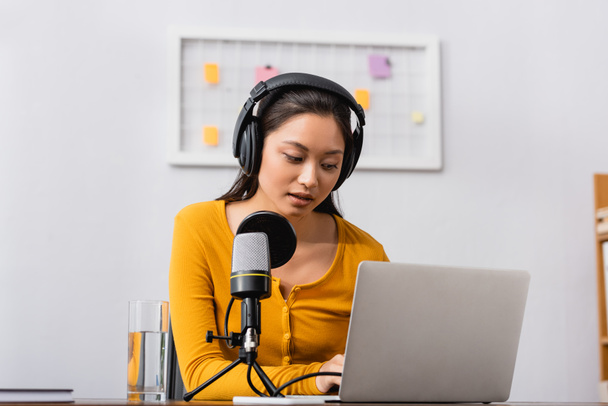 concentrado asiático radio host en inalámbrico auriculares usando portátil cerca de micrófono en estudio - Foto, Imagen