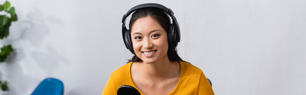 horizontaal beeld van vrolijke Aziatische omroep in draadloze hoofdtelefoon kijkend naar camera in radiostudio - Foto, afbeelding