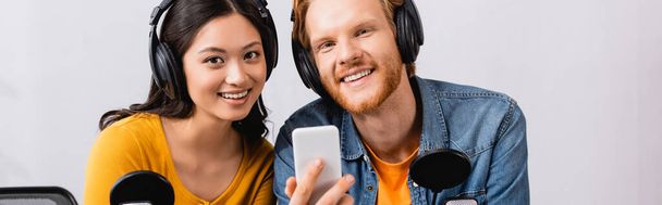 panoramic concept of redhead radio host holding smartphone while looking at camera together with asian colleague - Photo, Image