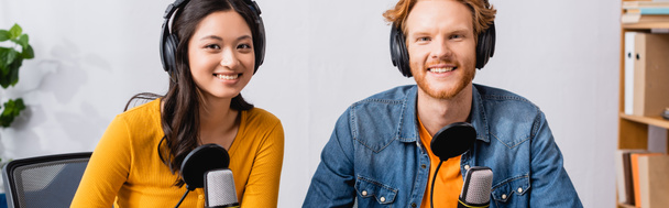 panoramic concept of excited interracial couple of broadcasters in wireless headphones looking at camera in radio studio - Photo, Image