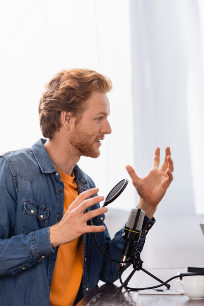 Kızıl saçlı, sakallı yayıncı iş yerinde mikrofonun yanında konuşurken el kol hareketi yapıyor. - Fotoğraf, Görsel