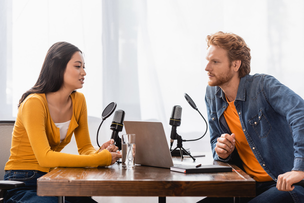 redhead radio host interviewing brunette asian woman near microphones and laptop in studio - Foto, imagen