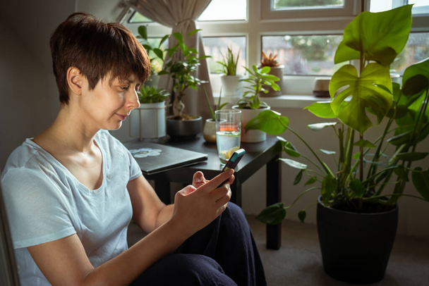 Close up mixed race woman in casual home clothes serfing in her smart phone sitting near coffee table with closed laptop and many green potted plants near the window. Working at home. Relaxing. - Foto, Bild
