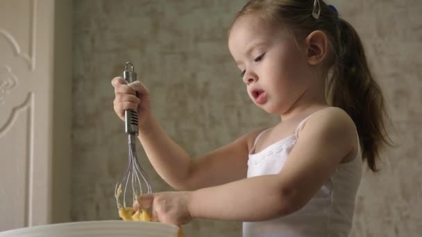Ein kleines Mädchen probiert rohen Teig mit Schneebesen und Peitschen. Kind bereitet Frühstück zu. Flüssiger hausgemachter Teig für Pfannkuchen und Pfannkuchen. Gesundes Frühstück für das Baby - Filmmaterial, Video