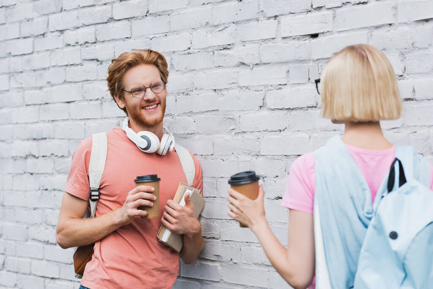 selective focus of bearded student looking at blonde friend while holding coffee to go near brick wall  - Photo, Image