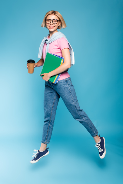 joven estudiante rubia sosteniendo taza de papel y cuadernos mientras vuela en azul - Foto, Imagen