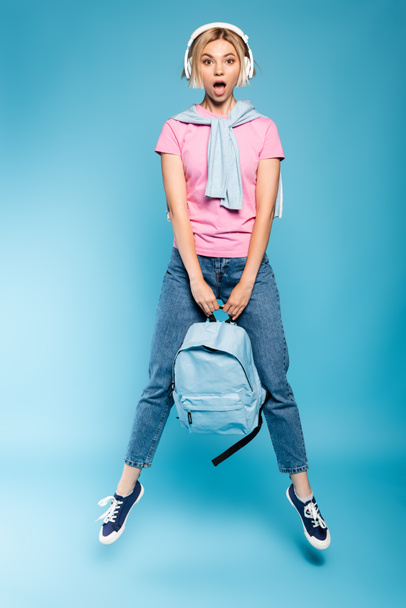 потрясенная блондинка в беспроводных наушниках смотрит в камеру, держа рюкзак и прыгая на синем  - Фото, изображение