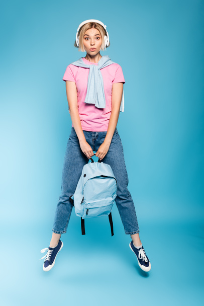 blonde student in wireless headphones looking at camera while holding backpack and jumping on blue  - Foto, Imagen