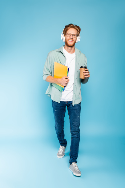 bearded student in glasses and wireless headphones holding paper cup and notebooks while walking on blue  - Foto, imagen