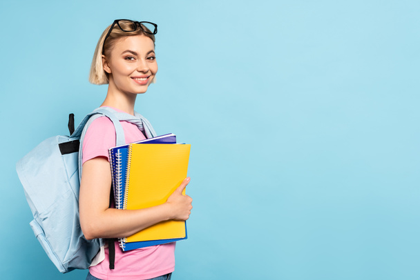 young blonde student with backpack holding notebooks on blue - 写真・画像