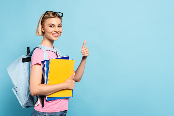 estudante loira com mochila segurando cadernos e mostrando o polegar no azul - Foto, Imagem