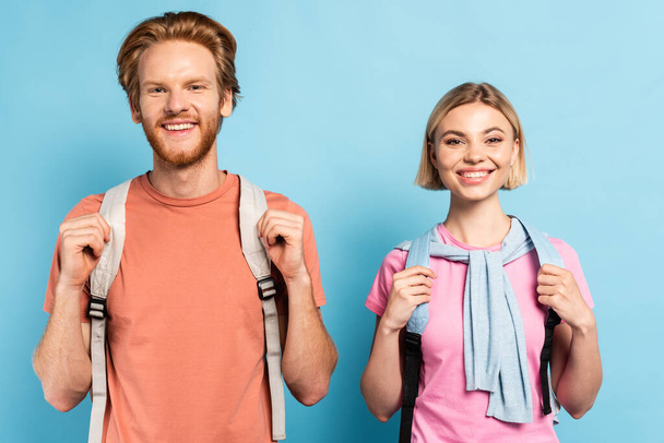 blonde and redhead students with backpacks looking at camera on blue - Фото, изображение
