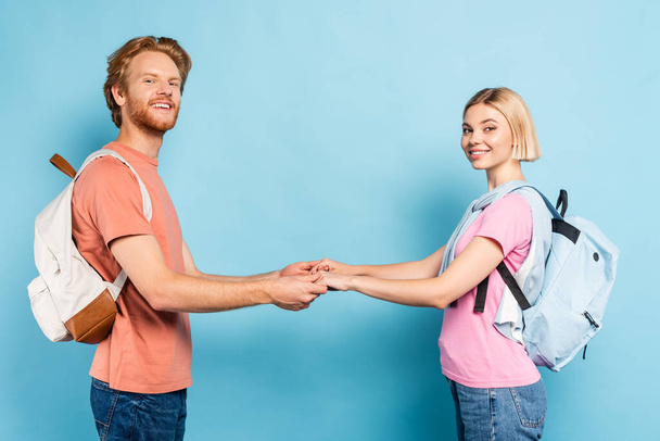 redhead and blonde students with backpacks looking at camera and holding hands on blue - 写真・画像