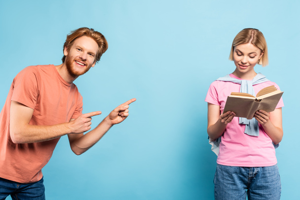 bearded student pointing with fingers at blonde woman reading book on blue  - Foto, Imagem