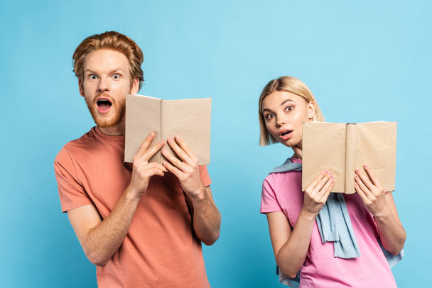 shocked redhead and blonde students holding books on blue - Фото, изображение