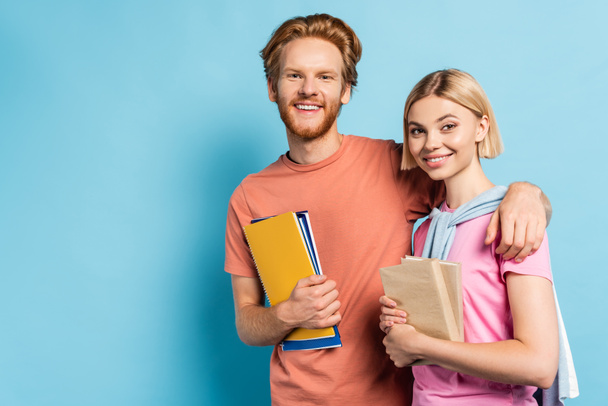 bearded man holding notebooks and hugging blonde student with books on blue - Photo, Image