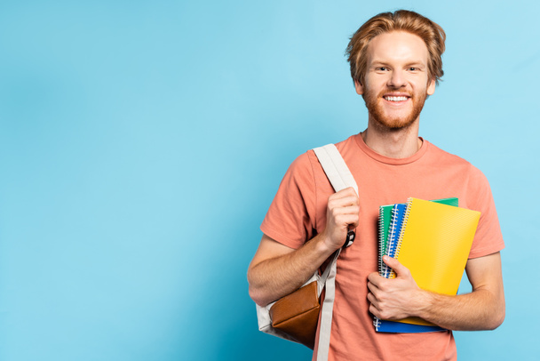 redhead student holding notebooks and touching backpack on blue - Fotó, kép