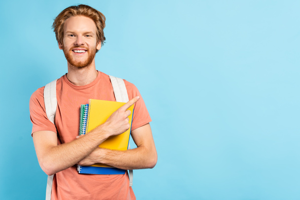 redhead student holding notebooks and pointing with finger on blue - Foto, Bild