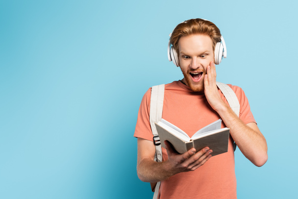 excited and redhead student in wireless headphones reading book on blue  - Foto, Imagem