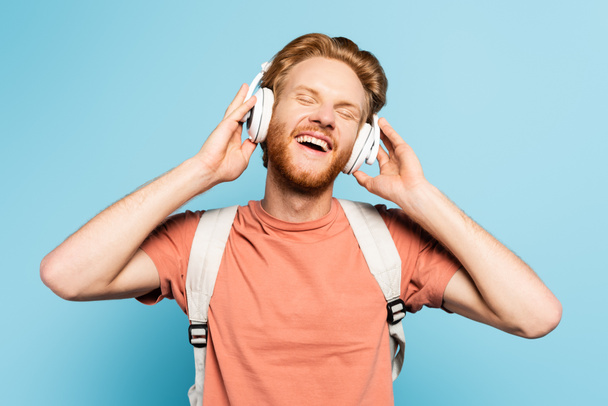 estudiante pelirroja con los ojos cerrados tocando auriculares inalámbricos en azul  - Foto, Imagen