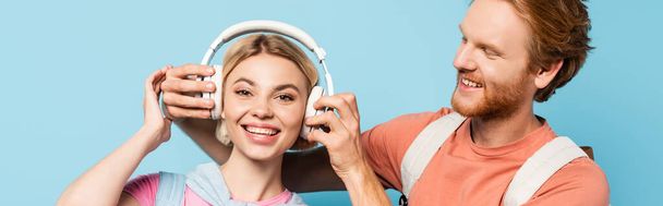 panoramic shot of bearded student touching wireless headphones of blonde friend on blue  - Фото, изображение