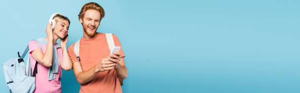 imagen horizontal de mujer rubia escuchando música en auriculares inalámbricos mientras mira el teléfono inteligente en manos de un estudiante en azul - Foto, imagen