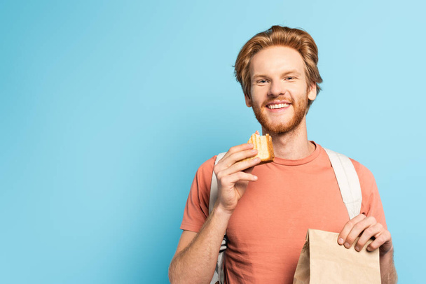young redhead student holding paper bag and toast bread on blue  - Photo, Image