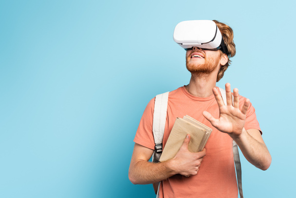 young redhead student in virtual reality headset holding books and gesturing on blue - Foto, Imagen