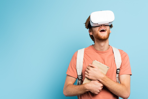 excited redhead student in virtual reality headset holding books and gesturing on blue - Foto, immagini