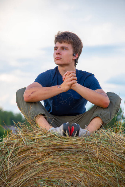Russia, Moscow region, August 2020. A handsome young man is sitting on a haystack in the fields against a blue cloudy sky. - Foto, Bild