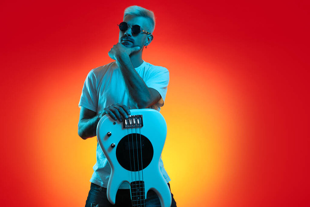 Blonde stylish hipster man holding posing with bass guitar in neon lights. Rock music concept. - Foto, Bild