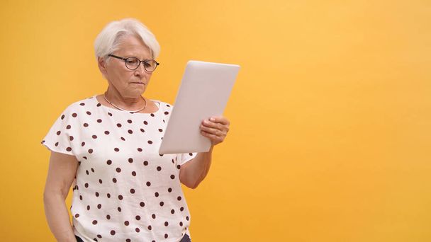 Una signora anziana confusa con un tablet regalo che si guardava intorno in cerca di aiuto. Isolato su sfondo arancione - Foto, immagini