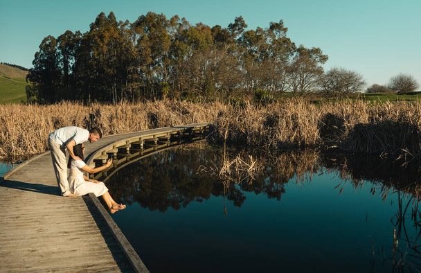 pareja joven sentada en el paseo marítimo en la orilla del lago y mirando el teléfono móvil. Peka Humedales de Peka, Hawke 's Bay, Nueva Zelanda. - Foto, imagen