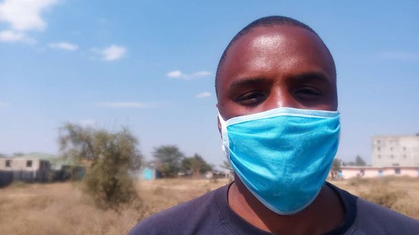 Közelről afrikai férfi kék sebészeti maszkot visel. Az orra és a szája el van takarva. A kamera felé néz. Napsütéses nap. Háttér homályos a föld, épületek és az ég.Coronavirus koncepció. - Fotó, kép
