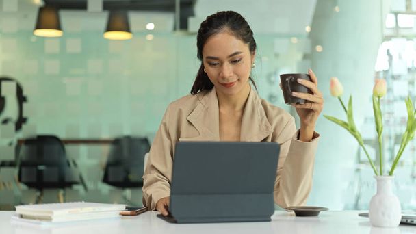 Portré női vállalkozó kezében egy csésze italt, miközben dolgozik a digitális tabletta irodai asztalon  - Fotó, kép