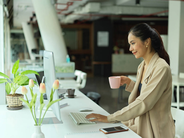 Ofisteki bilgisayar aygıtı üzerinde çalışırken bir yandan iş kadını kahve molası veriyor.  - Fotoğraf, Görsel