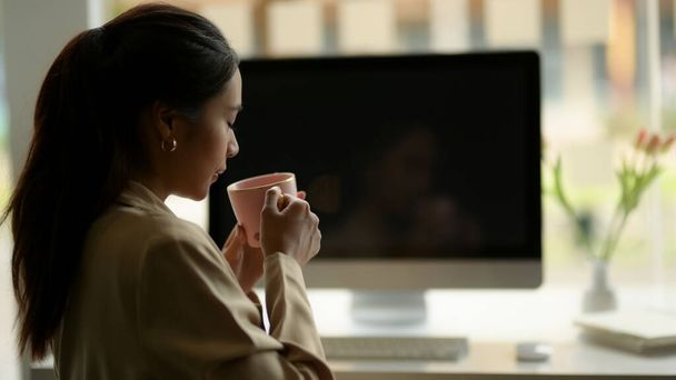 Портрет сотрудницы офиса расслабленной сидеть и пить кофе за компьютерным столом в офисной комнате  - Фото, изображение