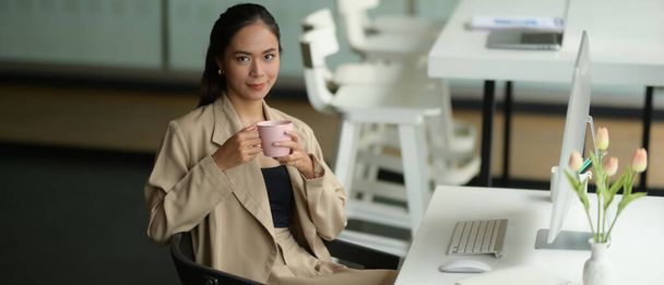Portrait of female office worker smiling and looking into camera while relaxing with a cup of beverage at workplace - Foto, Imagem