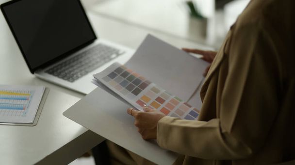 Imagen recortada de la diseñadora femenina eligiendo el color mientras trabaja con el ordenador portátil en la mesa de reuniones - Foto, Imagen
