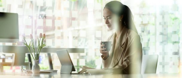 Vista lateral de una trabajadora de oficina sosteniendo una taza de café mientras trabaja con una tableta digital en una mesa blanca en un espacio de trabajo compartido - Foto, Imagen